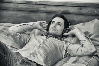 Handsome young man in bed listening to music with headphones, using cell phone or MP3 player