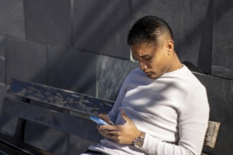 Male person sitting on a bench looking at his mobile phone, horizontal close up