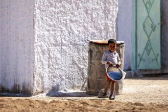 A child stands with a bucket in the sun next to a white house wall, Little boy stands by the house