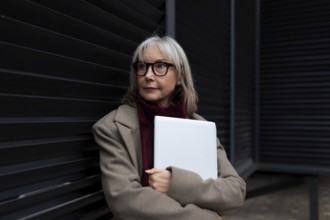 A middle-aged businesswoman with glasses is standing against a dark wall, holding a laptop. She