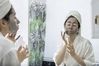 Portrait of a young woman applying a black cream face mask for skin in front of her bathroom mirror