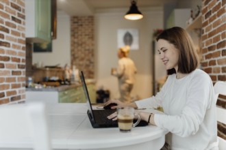 Woman happily using a laptop in a kitchen, enjoying her work with a relaxed demeanor