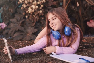 Blond kid girl selfie photo with tablet pc lying on grass turf with headphones