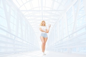 Beautiful young woman with music headphones standing on the bridge with a takeaway coffee mug and