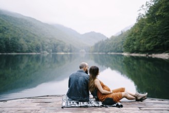 Man with pregnant woman are sitting on a pier by the lake in the park.