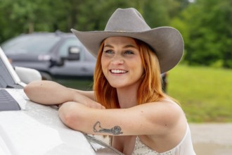 A beautiful redhead model poses in a country setting wearing cowgirl attire against her truck