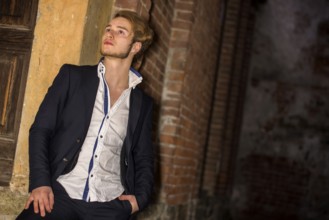 Handsome blond young man standing against brick wall, looking up to a side, wearing white shirt and