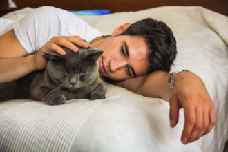 Handsome Young Animal-Lover Man on a Bed, Hugging and Cuddling his Gray Domestic Cat Pet