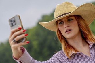 A beautiful redhead model uses her cellphone while posing in a country setting wearing cowgirl