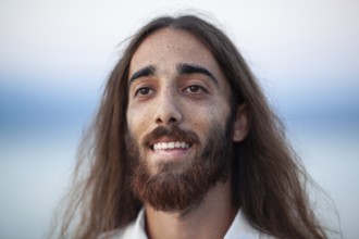 A portrait of a young man with long hair, a beard and a kind smile on his gace