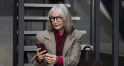 A middle-aged businesswoman dressed in a stylish coat and glasses is engaged with her smartphone