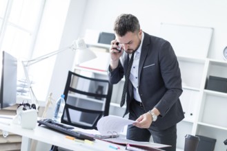 A man is standing near a table in the office, talking on the phone and flipping through documents