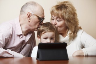 Grandparents kissing their little grandson who is busy with touchpad