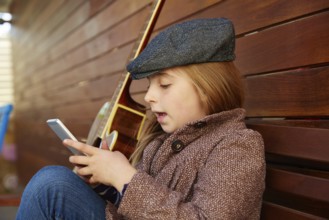 Blond kid girl playing smartphone winter beret and guitar on wooden background