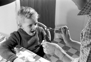 A boy is sick and won't take his medicine. His mother holds out a spoon and waits for him to gape