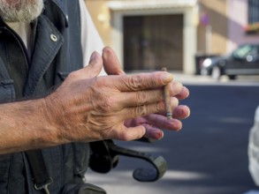 Older caucasian man sits on medical walker outdoors, smoking a cigar in public despite visible
