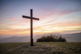 A cross stands in front of a colourful sunset in a peaceful landscape, Bisberg, Watterdingen,