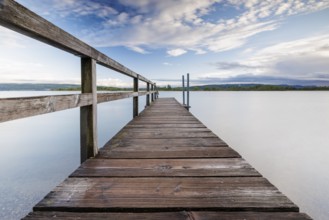 Long wooden footbridge leads into a quiet lake under a partly cloudy sky, Naturfreundehaus,