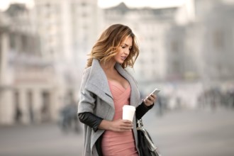 A businesswoman checking emails on her mobile phone and holding a coffee cup in her hand in front