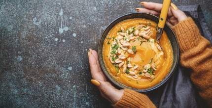Female hands in yellow knitted sweater holding a bowl with pumpkin cream soup on dark stone