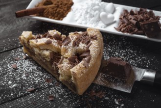Slice of apple pie with powdered sugar and chocolate, on a knife, on a black rustic table and a