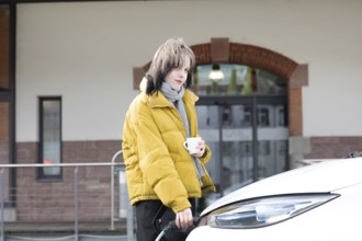 Woman in yellow jacket holding a cup, standing by charging electric car in urban environment,