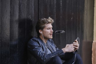 Young man listening music with smartphone earphones sitting in the street black door