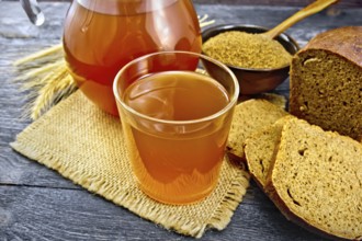 Kvass in glassful and glass jug on burlap, malt in a bowl, rye bread and ears on a wooden plank