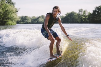 Young athletic man with long hair wakesurfing on waves of river in sunny summer weather. Ttheme