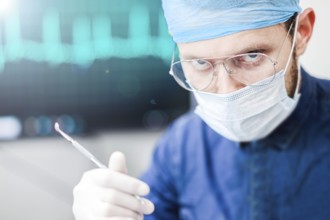 A serious surgeon with glasses and a mask, holding a tool in a medical setting