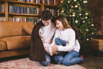 Happy family on Christmas Eve on the floor under a blanket near Christmas tree in the evening at