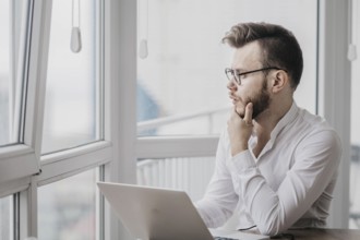 Pensive man in glasses and a white shirt looking out of a window beside his laptop