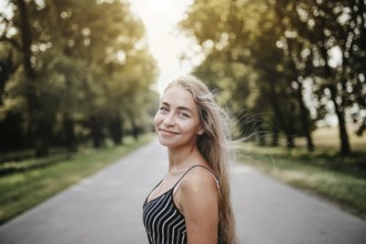 Side profile of a smiling woman on a road with trees, captured in a breezy atmosphere during sunset
