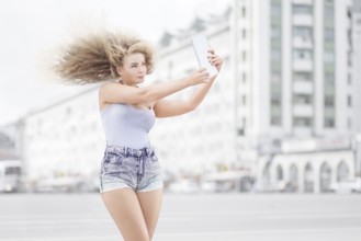 Happy young woman with vintage music headphones around neck taking selfie with tablet PC and