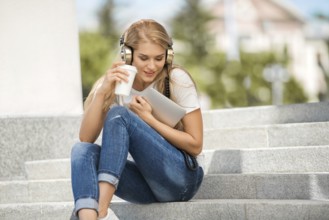 Happy young woman with vintage headphones, holding a takeaway coffee mug and a tablet PC, sitting