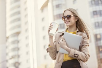 Beautiful young businesswoman with a disposable coffee cup, drinking coffee and holding a tablet in