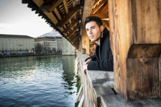 Young dark haired man standing on wooden medieval footbridge in Switzerland while looking away
