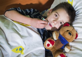 Sick child in bed with teddy bear. Measuring the temperature with a thermometer