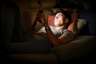 Attractive Young Man Lying on Sofa at Night and Illuminated by Light from Screen of Cell Phone at