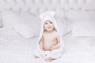 Adorable newborn baby boy with brown eyes sitting wrapped in a white towel with ears in light bed.