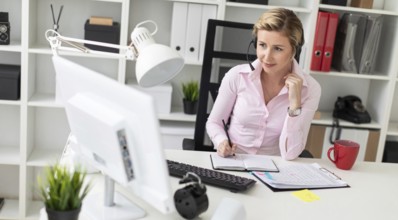 A young girl in headphones sits at a table in the office, holds a pencil in her hand and works with