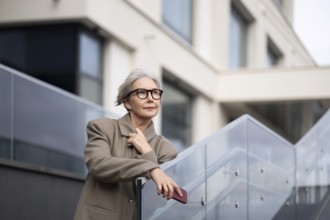 An older woman in fashionable glasses poses thoughtfully on a contemporary staircase, with a modern