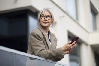 A senior businesswoman stands outside a contemporary building, holding her smartphone. She appears
