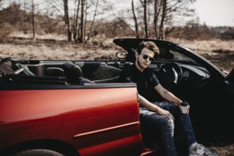 Man sitting in a red convertible, relaxed expression amidst a natural background
