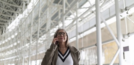 A middle-aged businesswoman with glasses is engaged in a phone conversation while strolling through