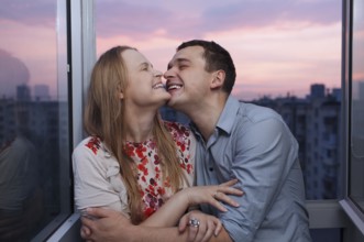 Playful young couple embracing and laughing on the balcony at a high floor during sunset
