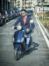 Young stylish man in suit and sunglasses driving scooter on empty road in sunny city