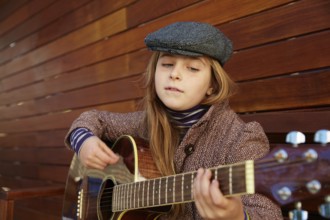 Blond kid girl playing guitar with winter beret and coat on wooden background