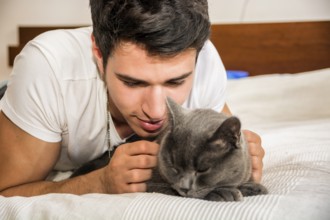 Handsome Young Animal-Lover Man on a Bed, Hugging and Cuddling his Gray Domestic Cat Pet