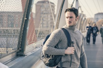 Handsome young man walking in city with backpack on one shoulder, wearing winter clothes